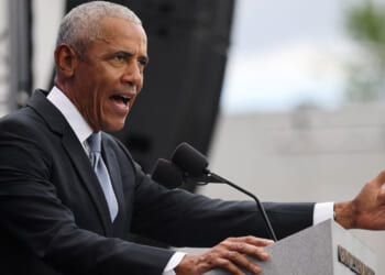 Barack Obama at a podium speaking with arm outstretched.