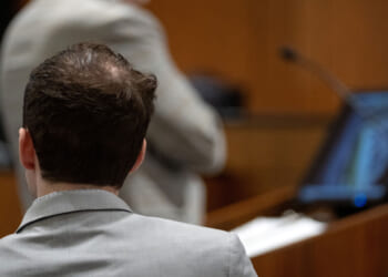 A view from behind of Tyler Robinson, in gray jacket, watching prosecutor present case against him.