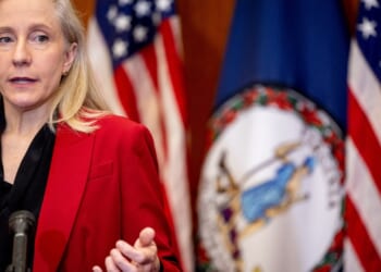 A woman in a red blazer speaks at a podium. Behind her are American flags and the Virginia state flag.