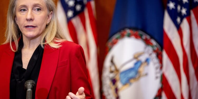 A woman in a red blazer speaks at a podium. Behind her are American flags and the Virginia state flag.