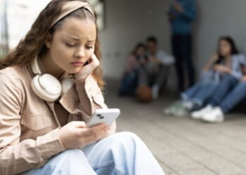 A young woman sits on a step on her phone outside a building on a bleak day ahead of a group of teens.