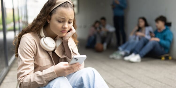 A young woman sits on a step on her phone outside a building on a bleak day ahead of a group of teens.