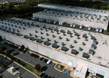 Aerial view of a large data center with rows of rooftop cooling units, surrounded by parking lots and greenery.