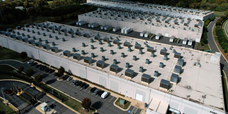 Aerial view of a large data center with rows of rooftop cooling units, surrounded by parking lots and greenery.