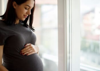 A woman rubs her stomach bulge near a window.