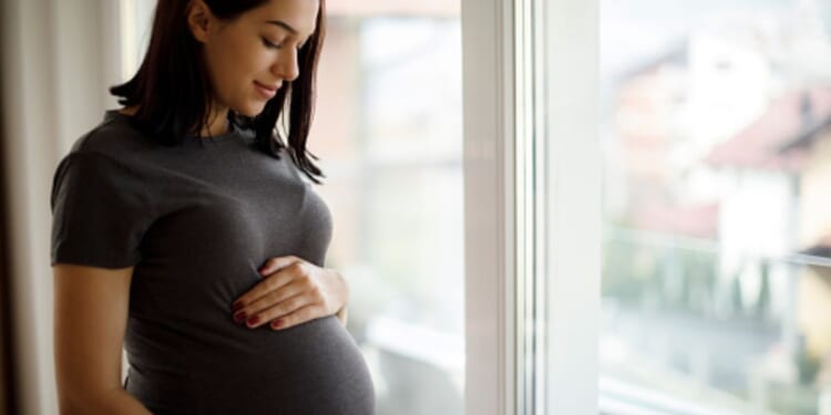 A woman rubs her stomach bulge near a window.