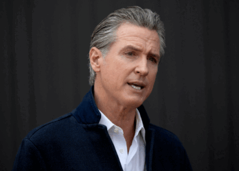 Governor Gavin Newsom, wearing a white shirt and dark jacket, speaks in front of a plain, dark background.