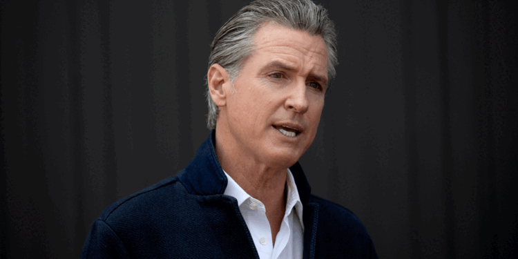 Governor Gavin Newsom, wearing a white shirt and dark jacket, speaks in front of a plain, dark background.