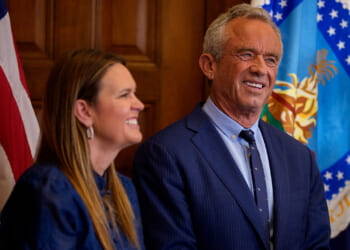 RFK Jr. in a blue suit and Gov. Sanders in a blue dress stand together indoors in front of an American flag.