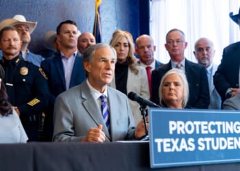 A group of people, including officials and police, sit and stand behind a table with a sign that reads Protecting Texas Students. One man at the center speaks into microphones.