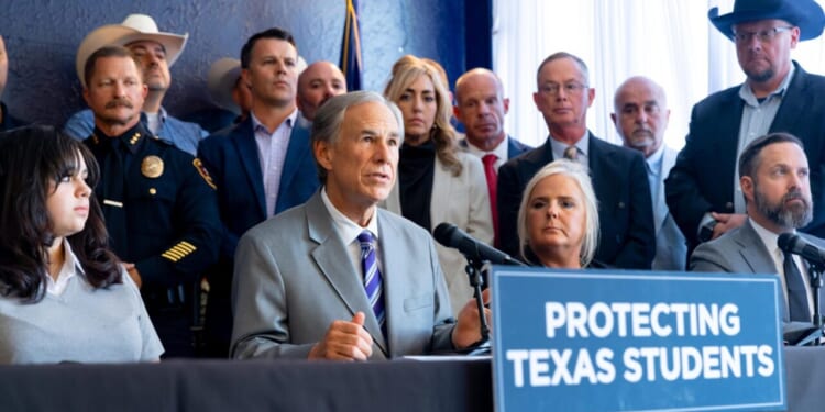 A group of people, including officials and police, sit and stand behind a table with a sign that reads Protecting Texas Students. One man at the center speaks into microphones.