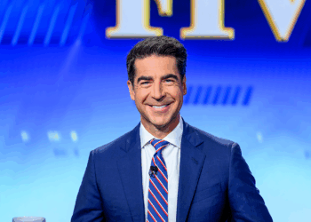 A man in a blue suit and striped tie smiles while seated in front of a blue background with large yellow and white letters partially visible behind him.