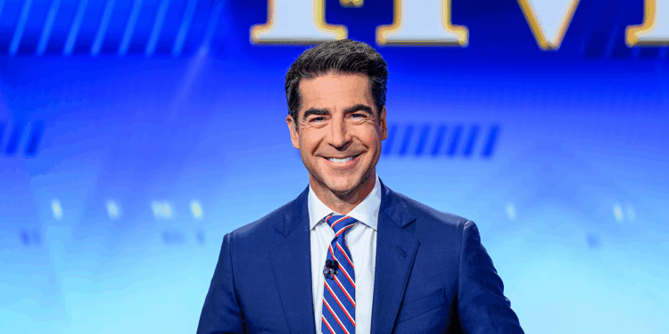 A man in a blue suit and striped tie smiles while seated in front of a blue background with large yellow and white letters partially visible behind him.