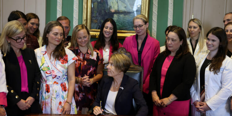 Massachusetts Gov. Maura Healey sits at a desk signing a document related to abortion, surrounded by a group of smiling women and men in professional attire, inside a formal room with a painting in the background.