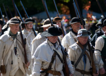 A group of people dressed in historical colonial clothing march outdoors, carrying long rifles upright over their shoulders. Trees with green and orange leaves are visible in the background.