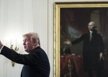 A man speaks into a microphone with his arm raised, standing in front of a large framed painting of another man in a historic setting.
