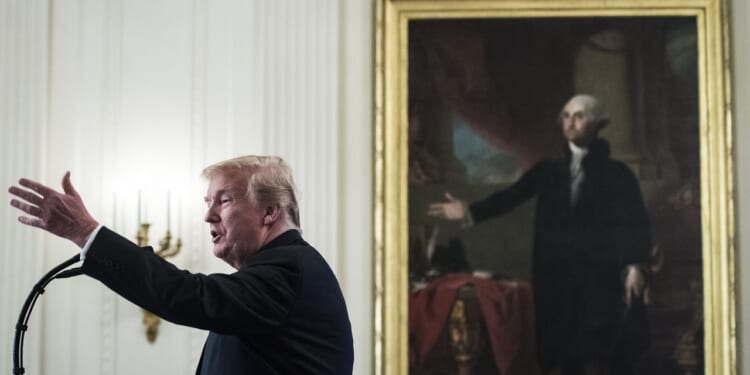 A man speaks into a microphone with his arm raised, standing in front of a large framed painting of another man in a historic setting.