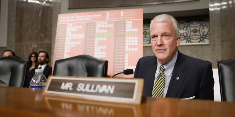 Sen. Dan Sullivan in a suit sits at a desk with a nameplate reading Mr. Sullivan. Behind him is a chart titled Russian & Chinese Incursions Into Alaska in a formal hearing room.