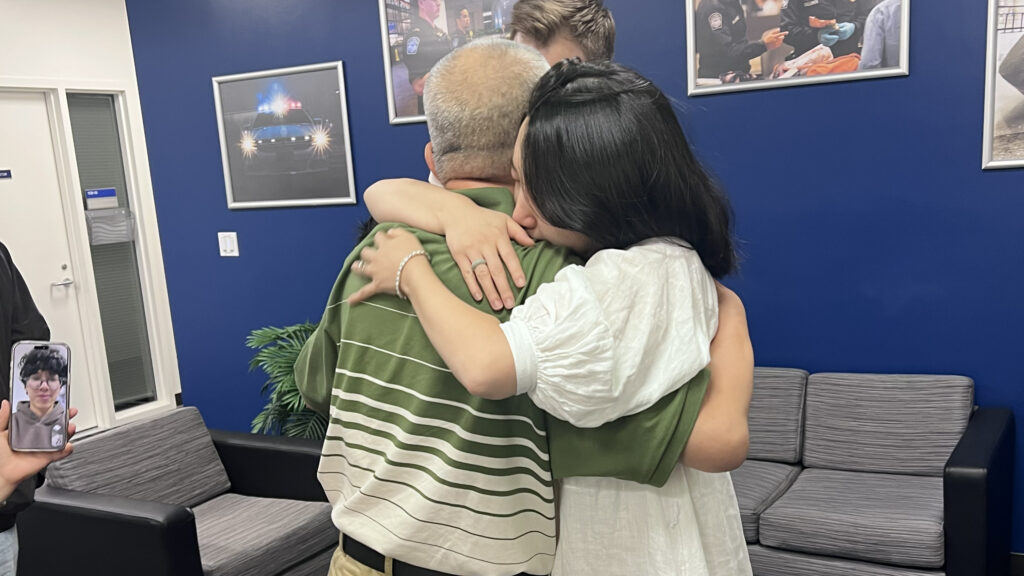 Three people embrace in a group hug in a room with blue walls and framed photos. One person holds a phone displaying a photo of someone—perhaps Jin. There is a gray couch and a potted plant in the background.