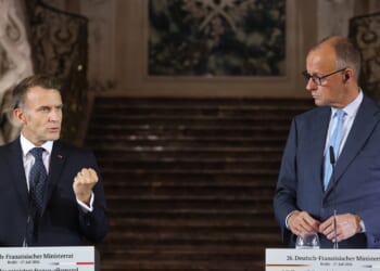Two men in suits stand at podiums, speaking at a formal event. One gestures with a closed fist while the other looks at him. Ornate stairs and decor appear in the background.