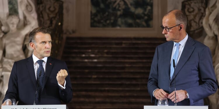 Two men in suits stand at podiums, speaking at a formal event. One gestures with a closed fist while the other looks at him. Ornate stairs and decor appear in the background.