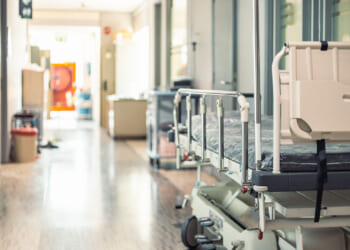 Empty hospital bed with raised side rails positioned in a bright, clean hallway, next to closed doors and medical equipment along the corridor walls.