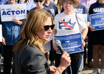 A woman in sunglasses speaks into a microphone at a rally. People behind her hold blue signs reading EDUCATORS FOR ACTION and DR. AMY ACTON FOR GOVERNOR. Some wear shirts supporting Amy Acton and Hambly.