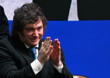 A man in a dark suit and blue tie is clapping and smiling, seated in front of a blue and black background.