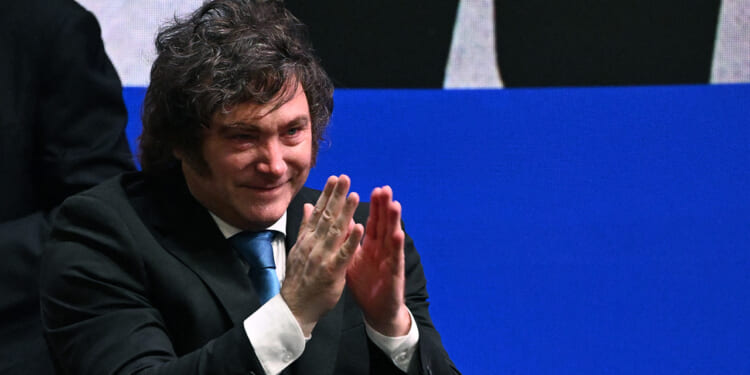 A man in a dark suit and blue tie is clapping and smiling, seated in front of a blue and black background.