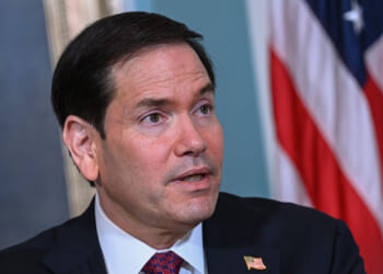 A man in a suit and tie with an American flag pin on his lapel sits in front of a U.S. flag, looking slightly upward with a neutral expression.