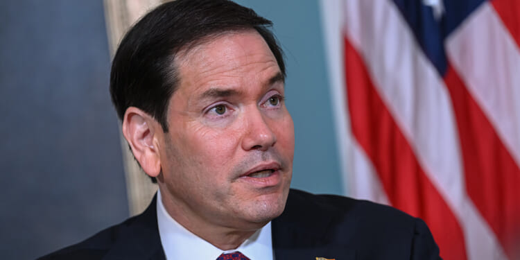 A man in a suit and tie with an American flag pin on his lapel sits in front of a U.S. flag, looking slightly upward with a neutral expression.