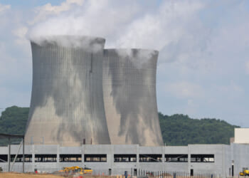 Two large concrete cooling towers emit white steam at a power plant, with green forested hills in the background and construction equipment visible in the foreground under a partly cloudy sky.