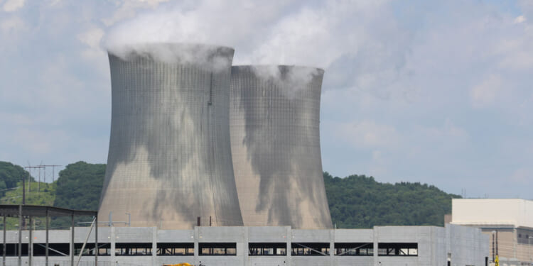 Two large concrete cooling towers emit white steam at a power plant, with green forested hills in the background and construction equipment visible in the foreground under a partly cloudy sky.