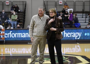 A man and a woman stand together on a basketball court, smiling. People are seated in the bleachers behind them, and a digital scoreboard displays the words Performance Therapy..