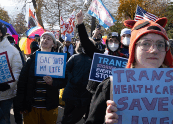 A group of people at a rally hold signs supporting transgender rights, including TRANS HEALTHCARE SAVES LIVES and GAC SAVED MY LIFE. Many wear hats, masks, and warm clothes; rainbow and trans flags are visible.