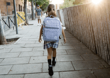 A young girl with a backpack walks on a city sidewalk in the morning sun. A man stands in the distance. Trees and buildings line the street.