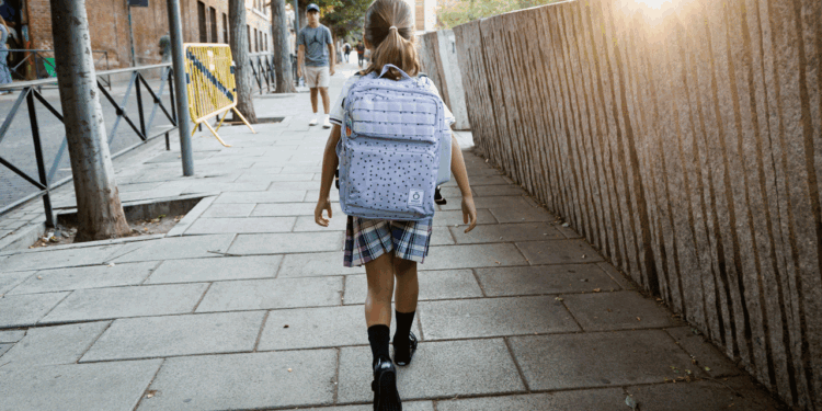A young girl with a backpack walks on a city sidewalk in the morning sun. A man stands in the distance. Trees and buildings line the street.