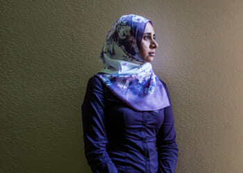 A woman wearing a floral hijab and a dark purple shirt stands against a plain beige wall, looking thoughtfully to the right.