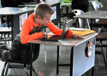 A young boy leans on his desk while using a laptop in a classroom. Papers and supplies are on the desk, and other empty desks are nearby.