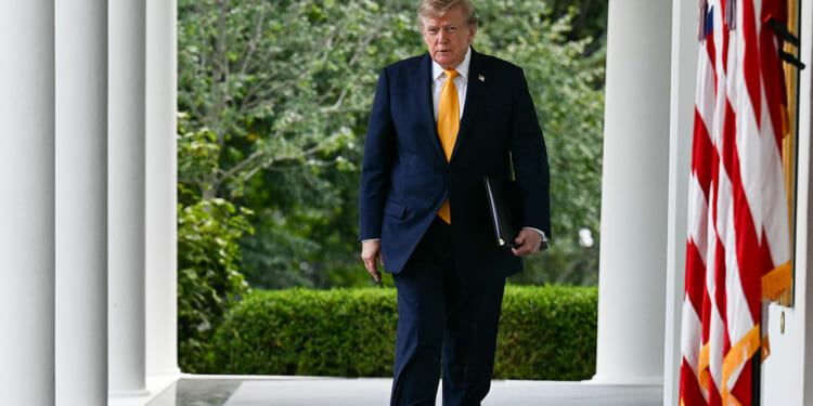 Donald Trump in a dark suit walks along a colonnade holding a folder. Greenery and trees are in the background. The U.S. flag is visible in the foreground on the right.