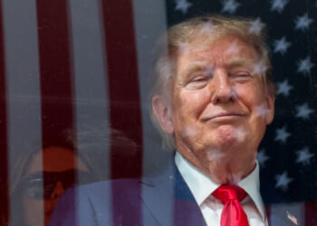 President Trump in a suit and red tie smiles while standing in front of a window with the reflection of the American flag. Another person with sunglasses is partially visible in the background.