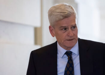 Sen. Bill Cassidy with short gray hair wearing a dark suit, white shirt, and patterned tie stands indoors, looking to his right with a neutral expression.