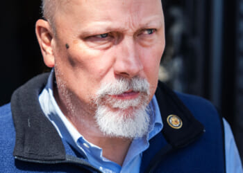 A bald man with a white goatee and a serious expression wears a blue vest and light blue shirt, standing outdoors with a dark background.