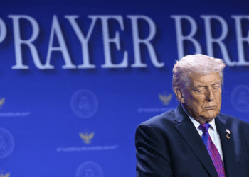A man in a suit stands with his eyes closed in front of a blue backdrop with the words PRAYER BREAK partially visible.