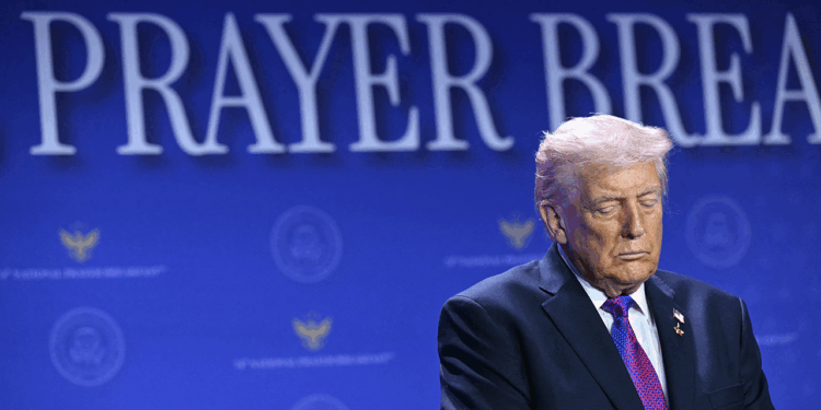 A man in a suit stands with his eyes closed in front of a blue backdrop with the words PRAYER BREAK partially visible.
