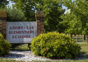 A brick sign reading Ashby-Lee Elementary School stands among green bushes and trees on a sunny day.