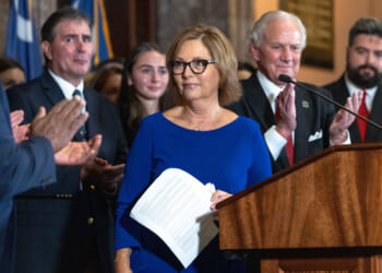 Darline Graham in a blue dress holding papers stands at a podium as people around her applaud. Several people in suits and formal attire are visible, and a flag is in the background.