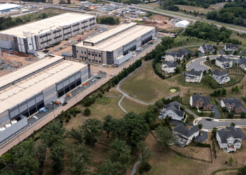 Aerial view showing two large industrial warehouse buildings next to a residential neighborhood with winding roads, single-family homes, trees, and a small pond. Construction equipment and materials are visible near the warehouses, which were recently developed as part of a local tax break initiative encouraging business growth in the area.
