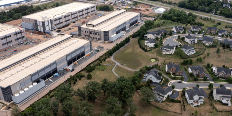 Aerial view showing two large industrial warehouse buildings next to a residential neighborhood with winding roads, single-family homes, trees, and a small pond. Construction equipment and materials are visible near the warehouses, which were recently developed as part of a local tax break initiative encouraging business growth in the area.