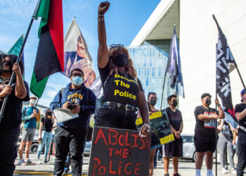 A group of people protest outdoors. One woman stands in front, raising her fist and holding a sign that says Abolish the Police. Many wear masks and hold flags.